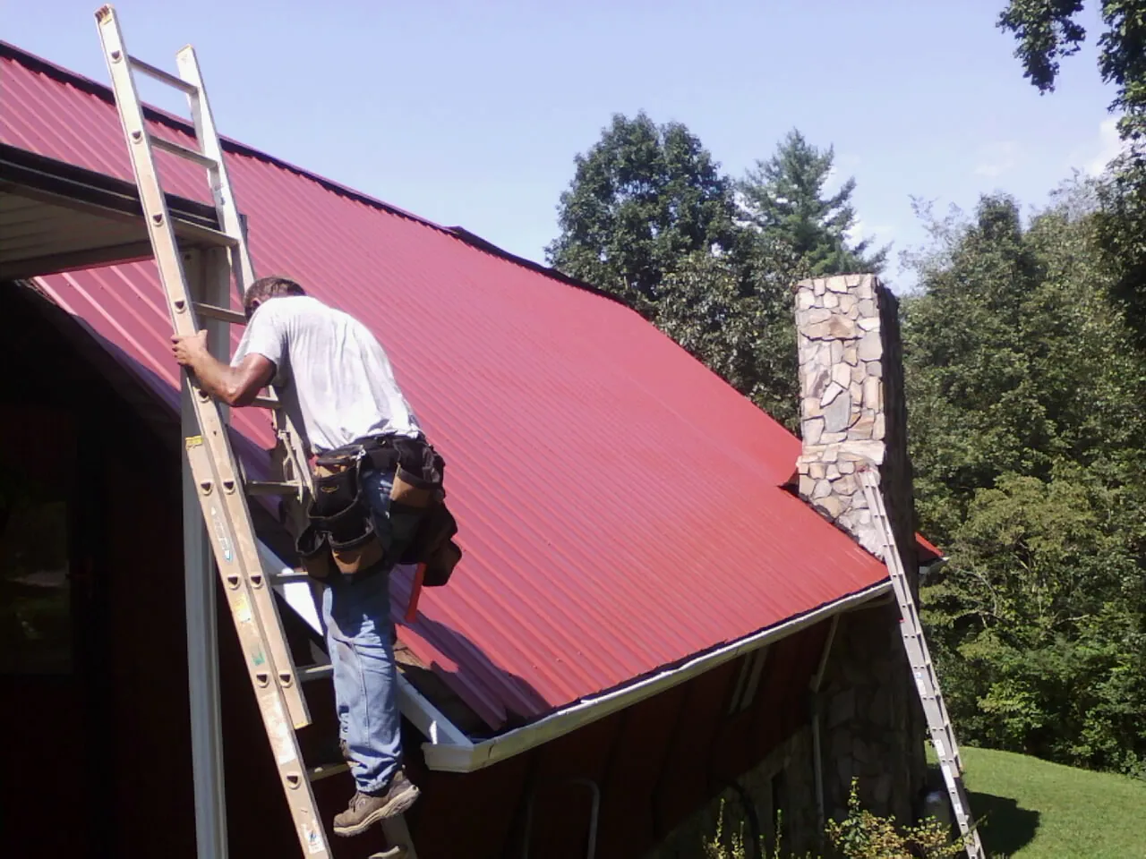 Roofer on a ladder beside a red metal roof and stone chimney — Soffit & Fascia Repair by Roofing Brothers in Sardinia